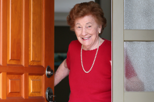 Elderly woman at home opening her front door to greet you with smile on her face