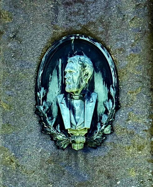 Sculpted bust of a distinguished man in a formal suit, framed by an ornate oval and surrounded by a laurel wreath, mounted on a textured gray stone wall.