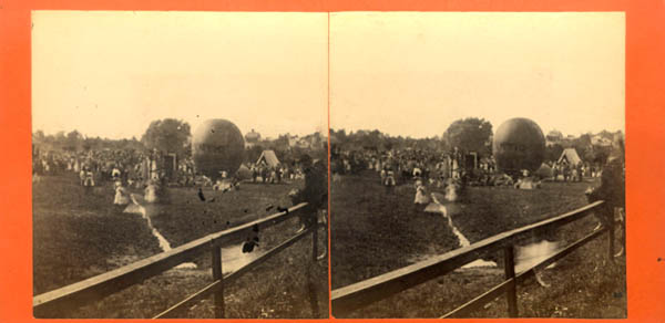 A historical photograph depicting a large outdoor crowd gathered for a ballooning event. Two large balloons are visible in the background, with people in period clothing nearby.